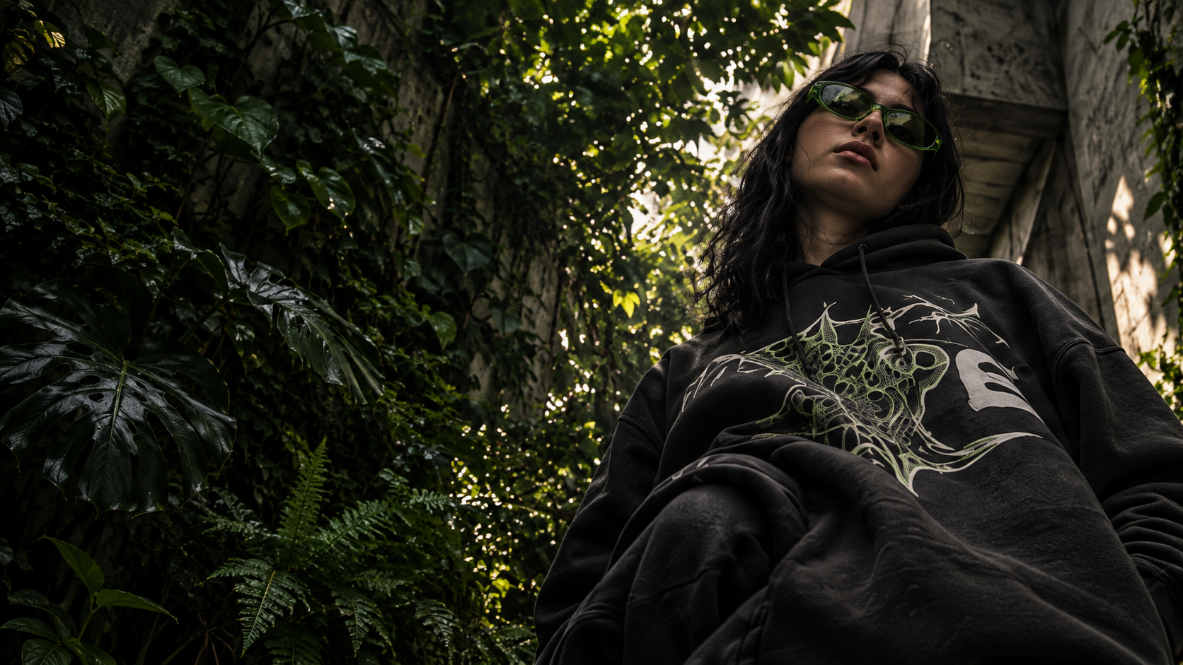 Modelo con gafas verdes y hoodie gráfico entre plantas y concreto.